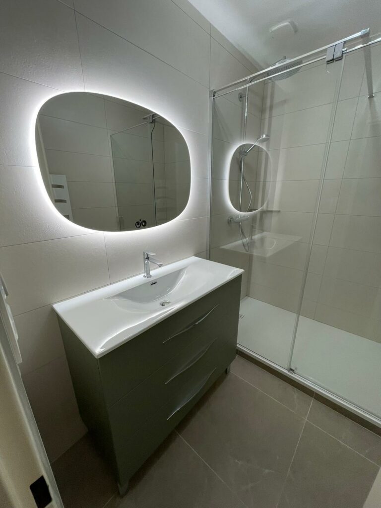 Salle de bain miroir arrondi au dessus de vasque blanche et maubleu sous vasque grise avec douche à l'italienne